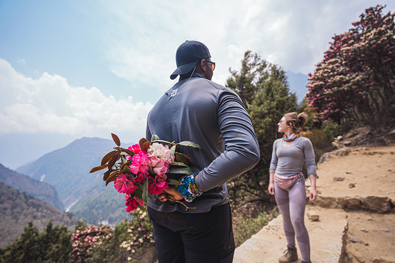 man holding flowers at his back and approaching a woman somewhere in the mountains